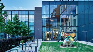 A modern building with large glass windows, outdoor seating area with chairs and tables, and a sculpture in the foreground. - Hyatt Regency Manchester (Manchester)