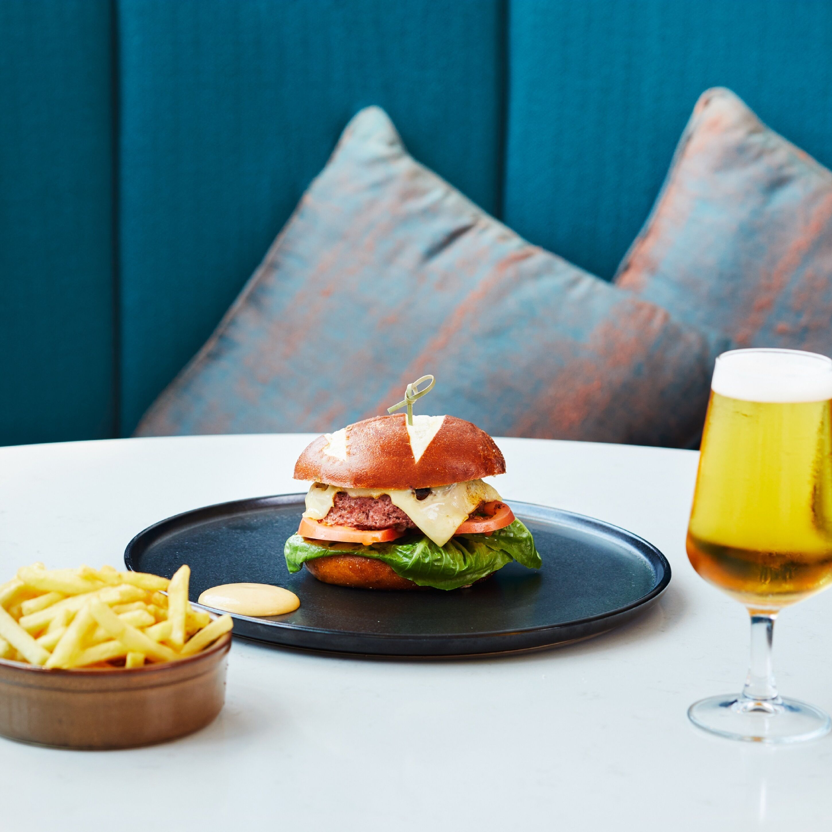 A burger on a black plate with fries and a glass of beer on a white table.