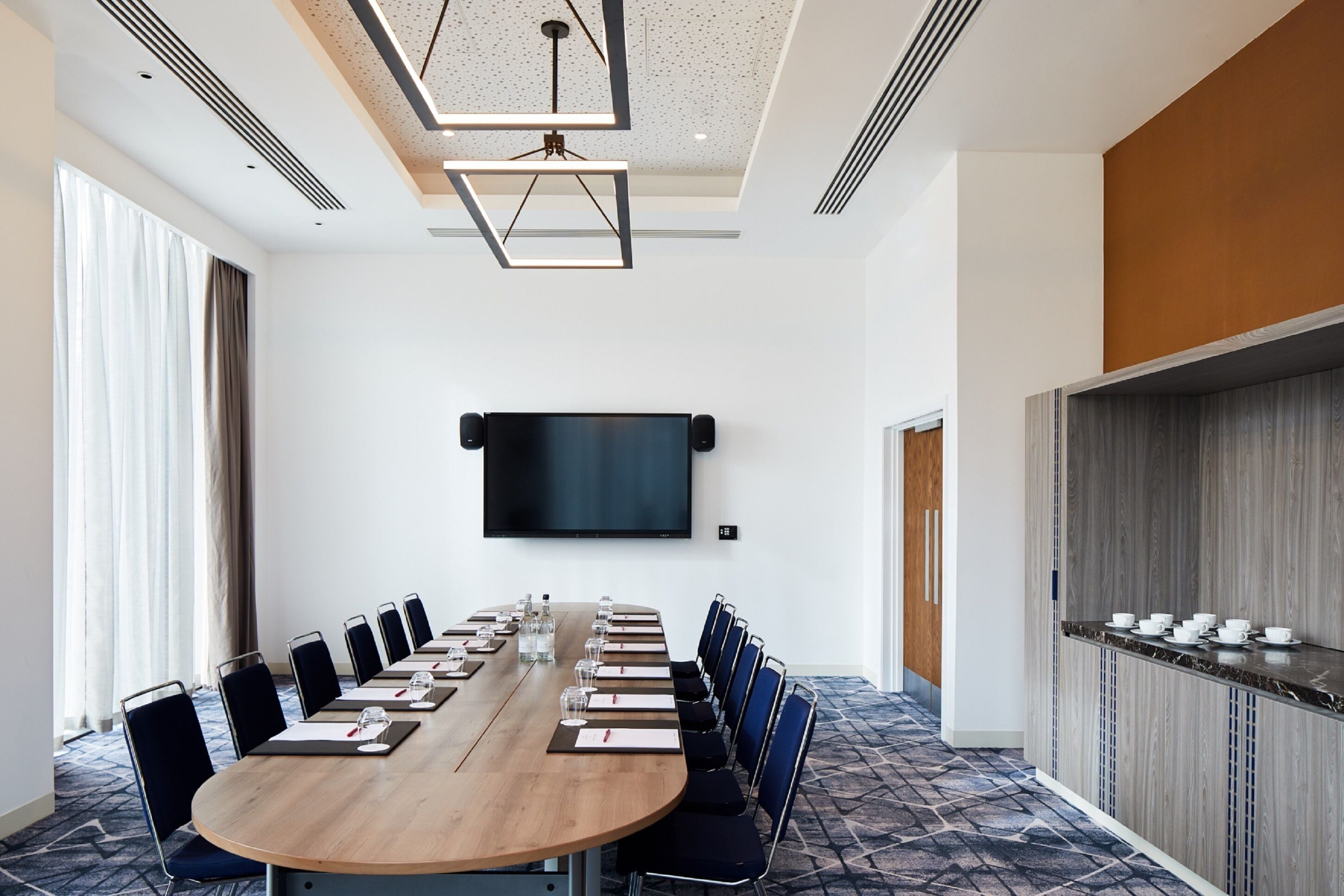A conference room with a long table, chairs, a flat-screen TV, and a bar area with cups and glasses.
