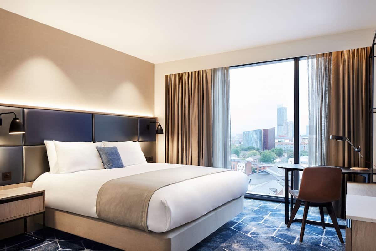 Ein Hotelzimmer mit einem großen Bett, einem Schreibtisch, einem Stuhl und Blick auf die Stadt durch große Fenster.