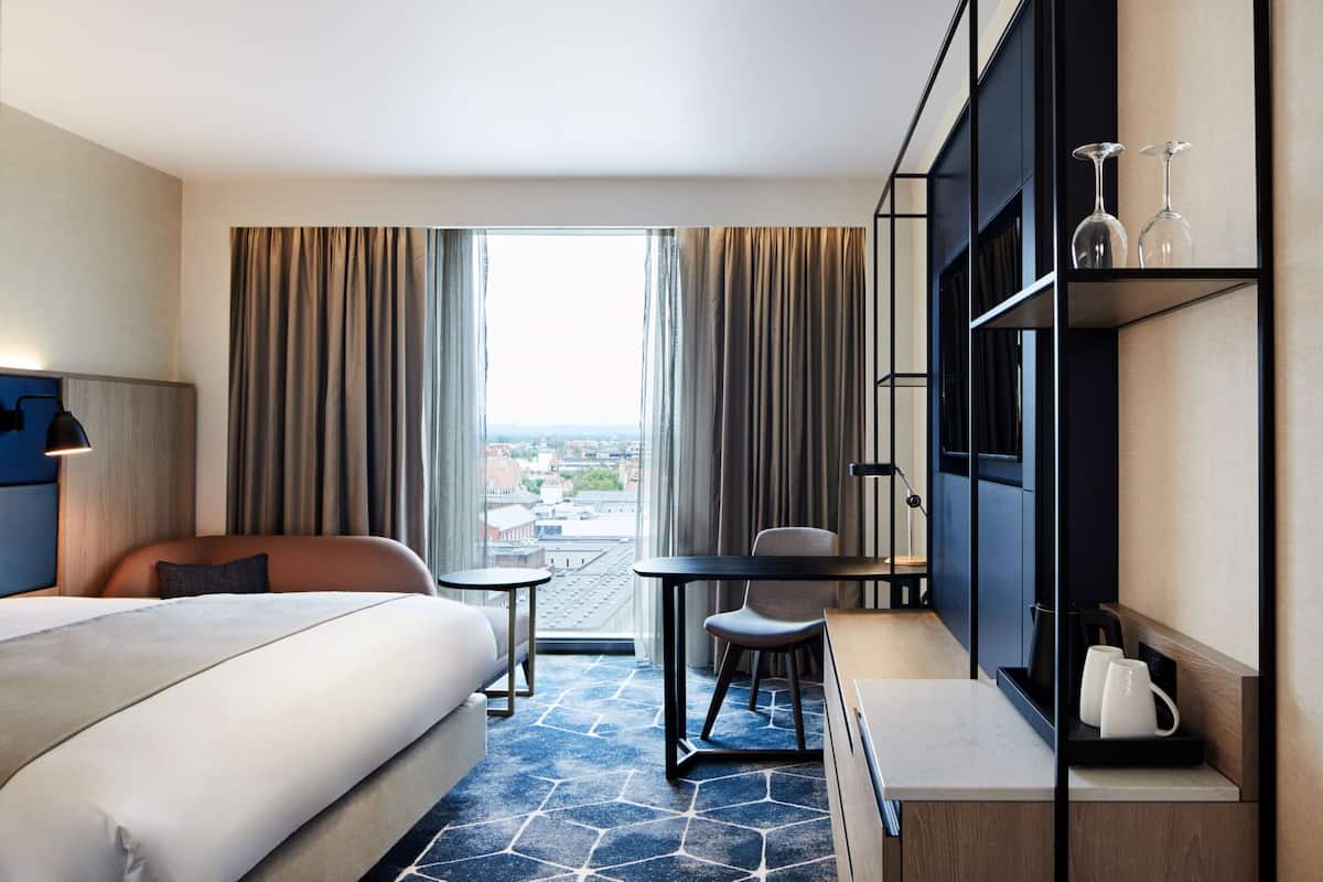 Ein Hotelzimmer mit einem großen Bett, einem Schreibtisch, einem Sessel, einem kleinen Tisch und einem Fernseher auf einem Regal.