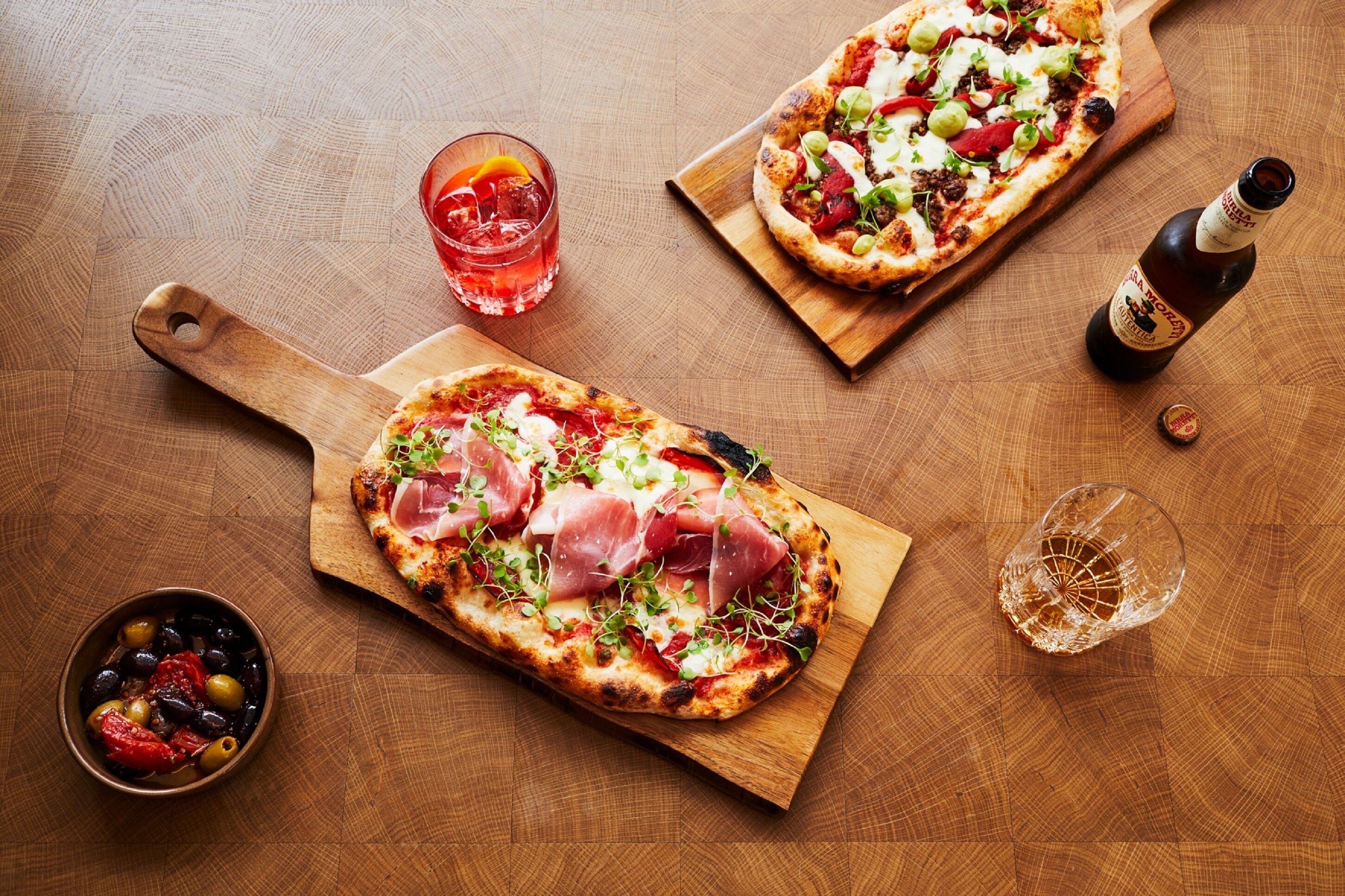 Two pizzas on wooden boards, a glass of red drink, a bottle of beer, and a bowl of olives.
