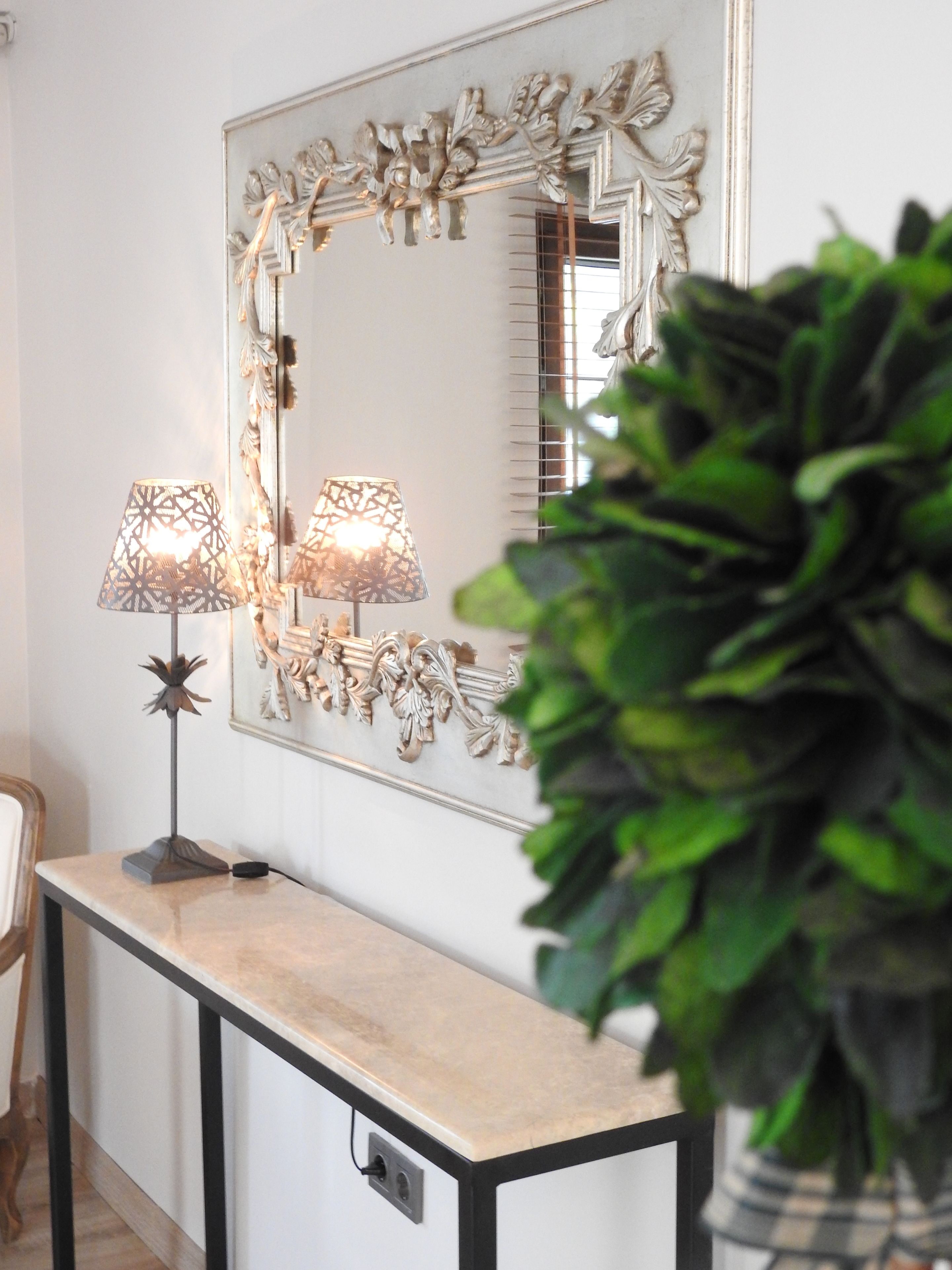 A room with a large ornate mirror, two lamps, a console table, and a potted plant.