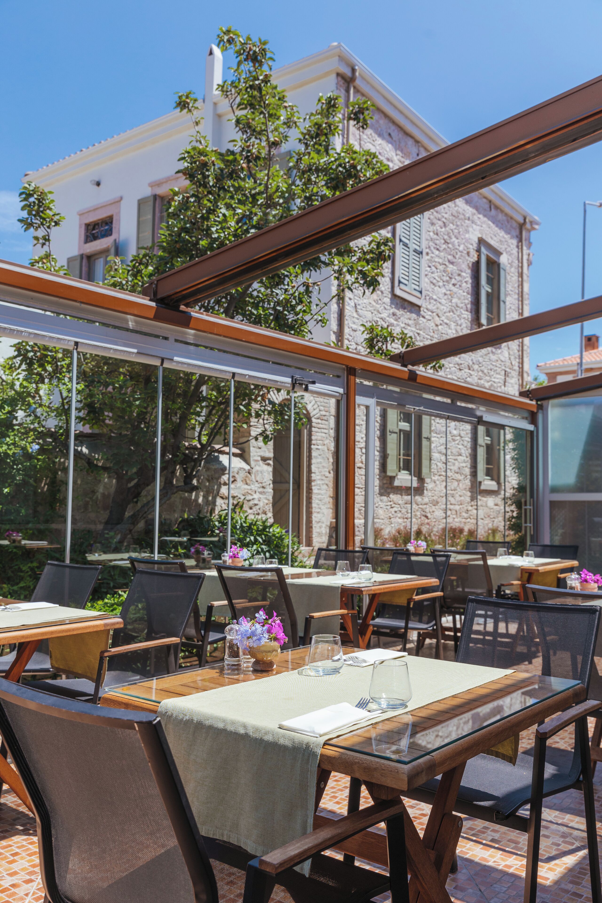 An outdoor dining area with tables and chairs, large windows, and a stone building in the background.