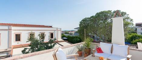 A rooftop terrace with a white sofa, wooden chairs, a table with fruit, and a potted plant.