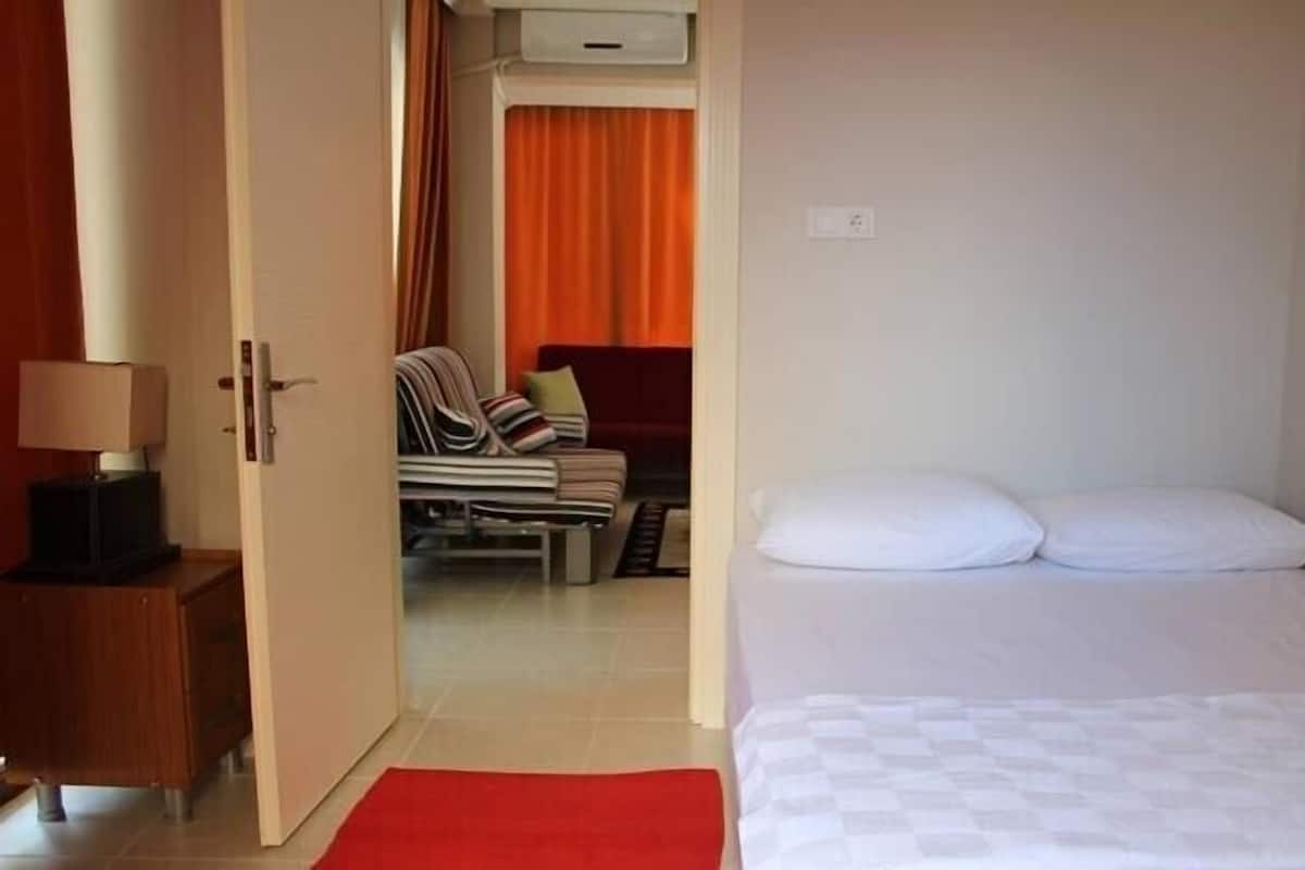 A hotel room with a bed, a nightstand, a red rug, and a view into the adjacent room with a chair and curtains.