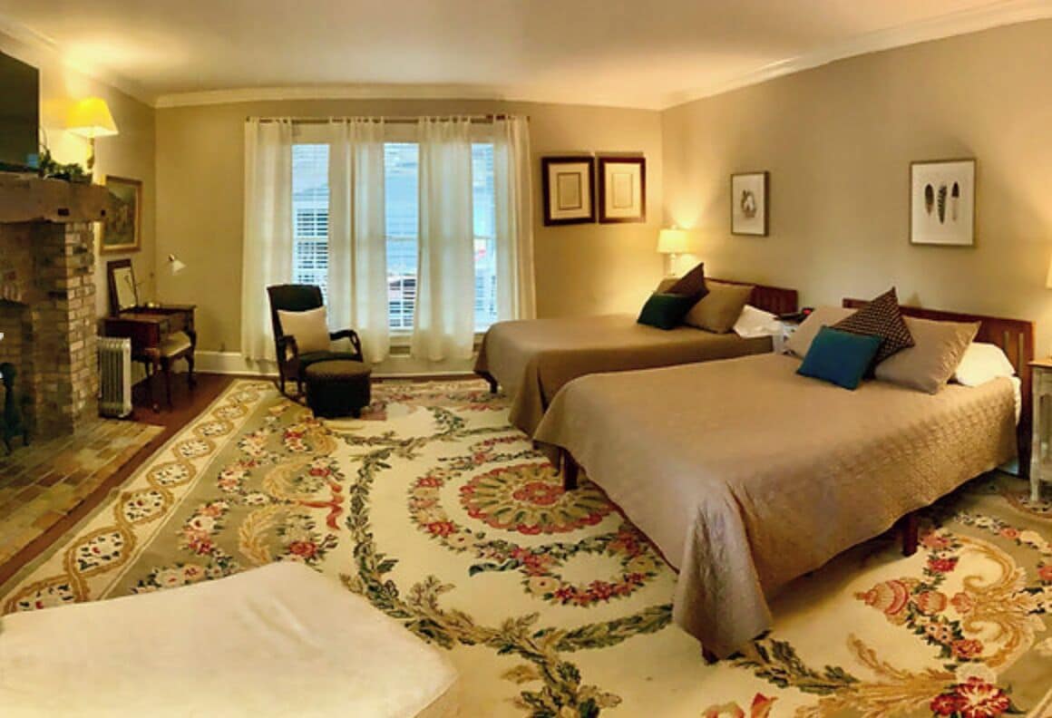 Une chambre d’hôtel avec deux lits, une cheminée, une grande fenêtre avec des rideaux et un sol recouvert de moquette.