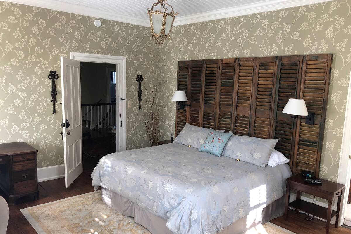 A bedroom with a large bed, wooden headboard, two bedside lamps, a nightstand, and a door leading to another room.