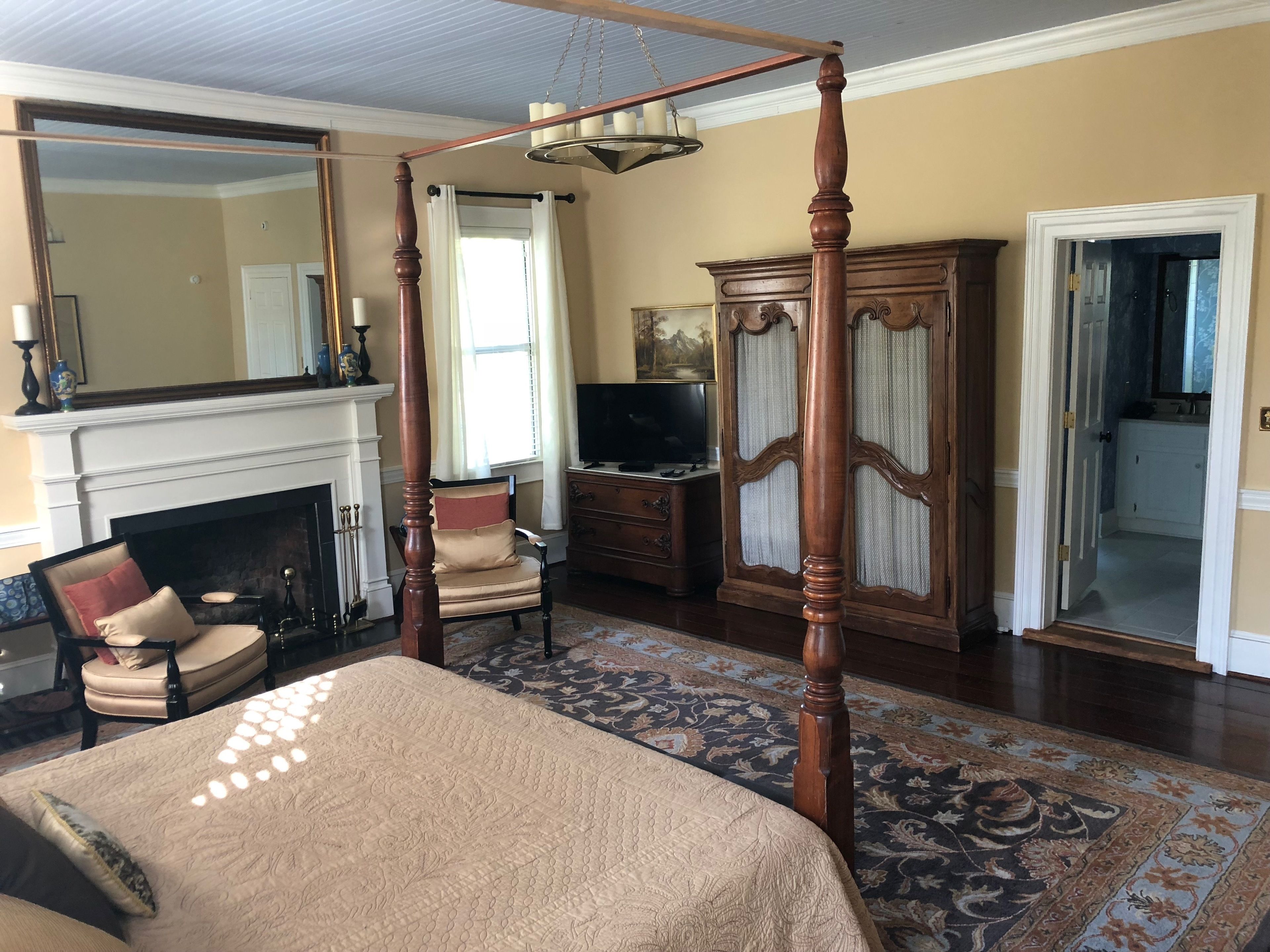 A four-poster bed, a fireplace, a television, and a large mirror in a room with a wooden floor and patterned rug.