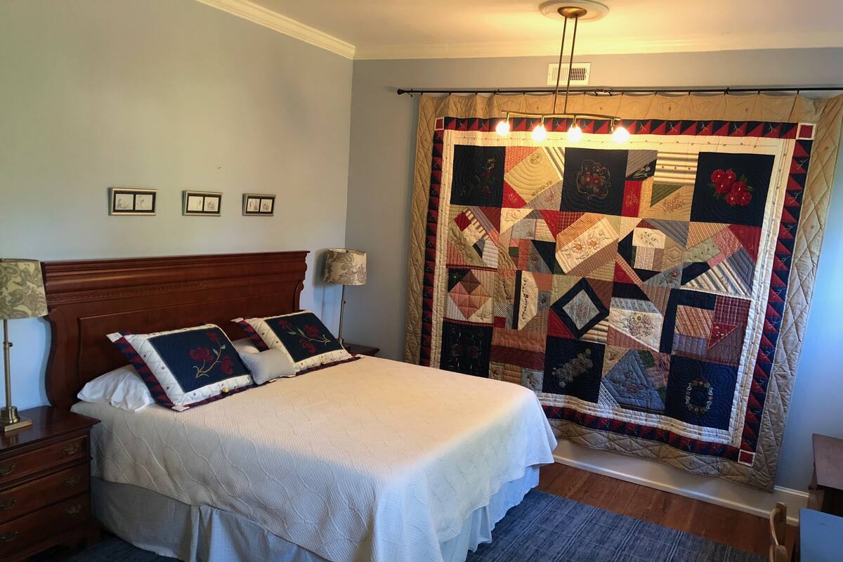 A bedroom with a large bed, a wooden headboard, a nightstand with a lamp, and a quilted curtain.