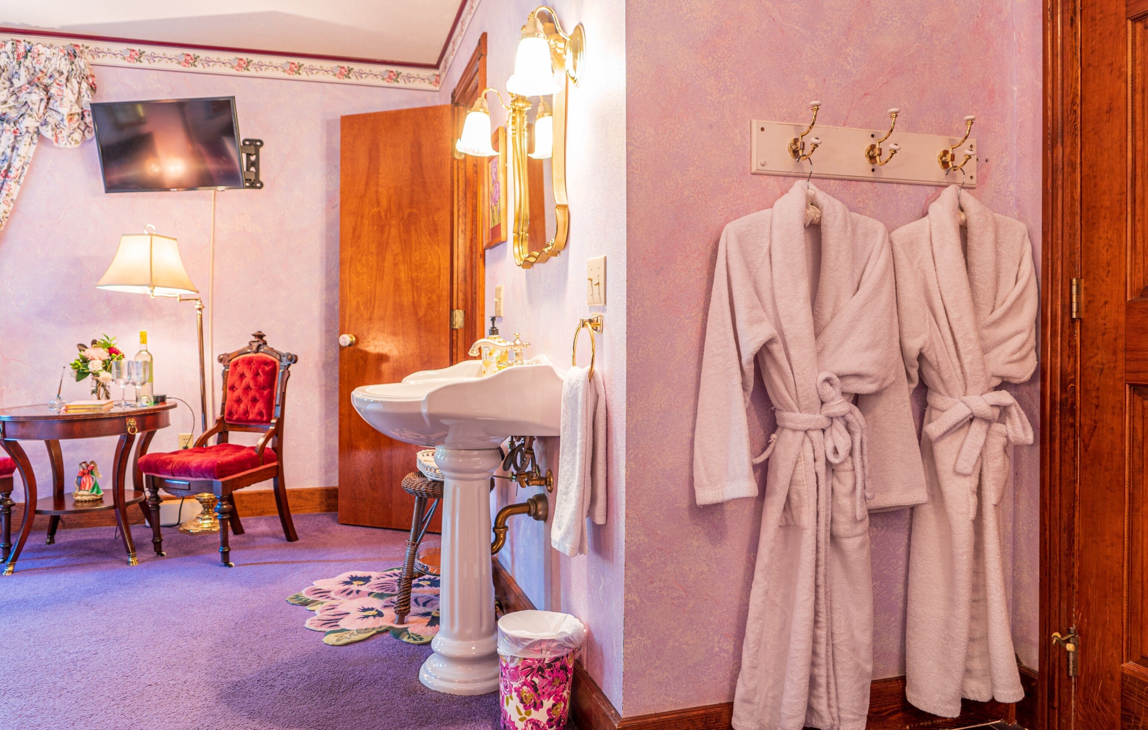 A bathroom with a pedestal sink, a red chair, a wooden door, and two white robes hanging on hooks.