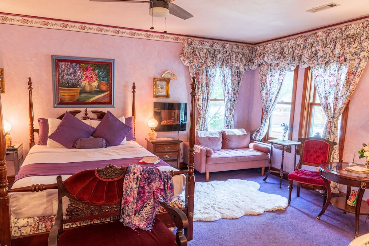 Un dormitorio con cama con dosel, una zona de estar con una silla, una mesita y una ventana con cortinas florales.