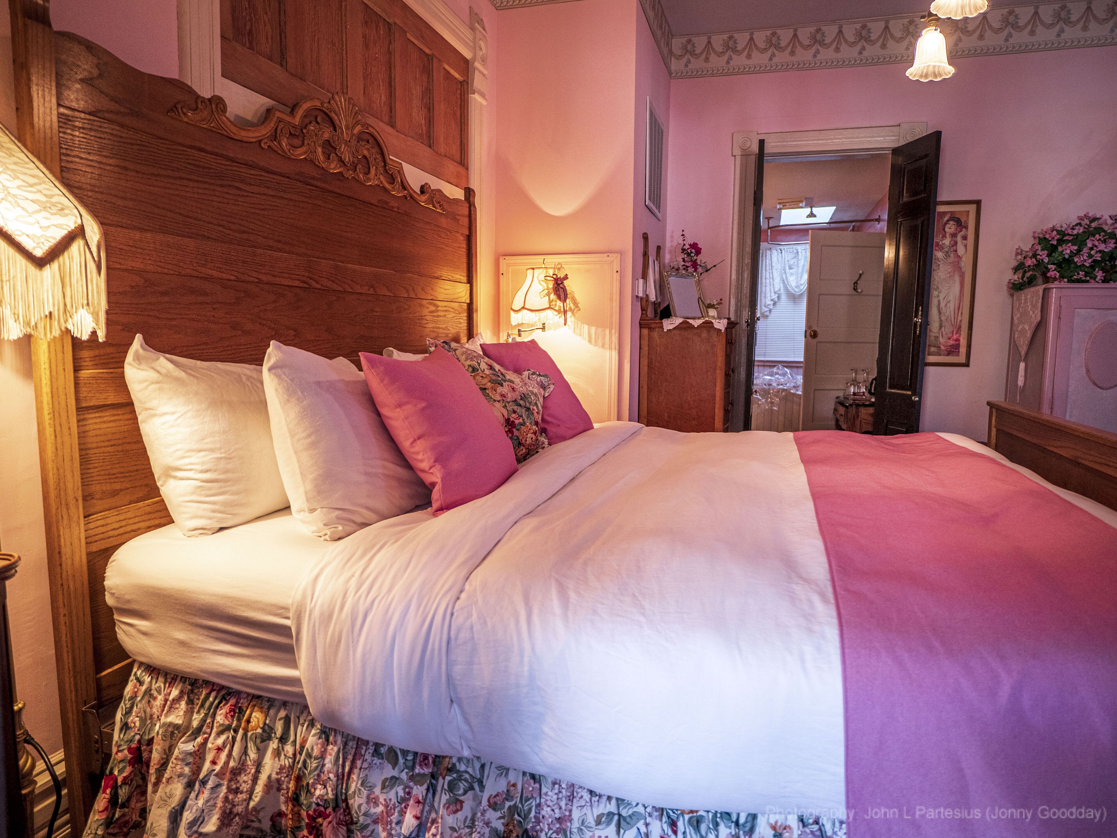 A neatly made bed with a wooden headboard, a floral skirt, and a pink blanket.