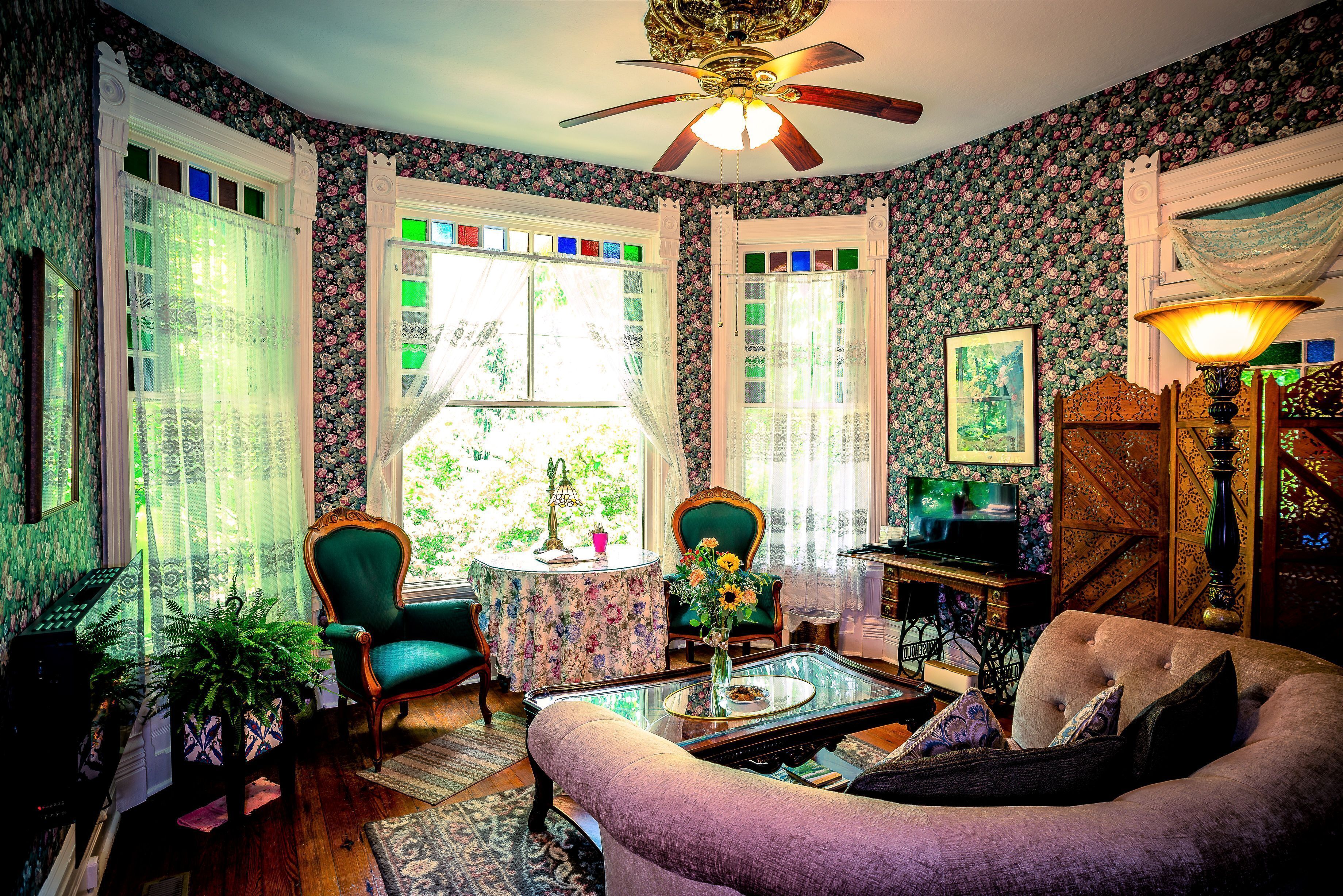 A room with a ceiling fan, floral wallpaper, a green armchair, a round table with a vase, and a television.