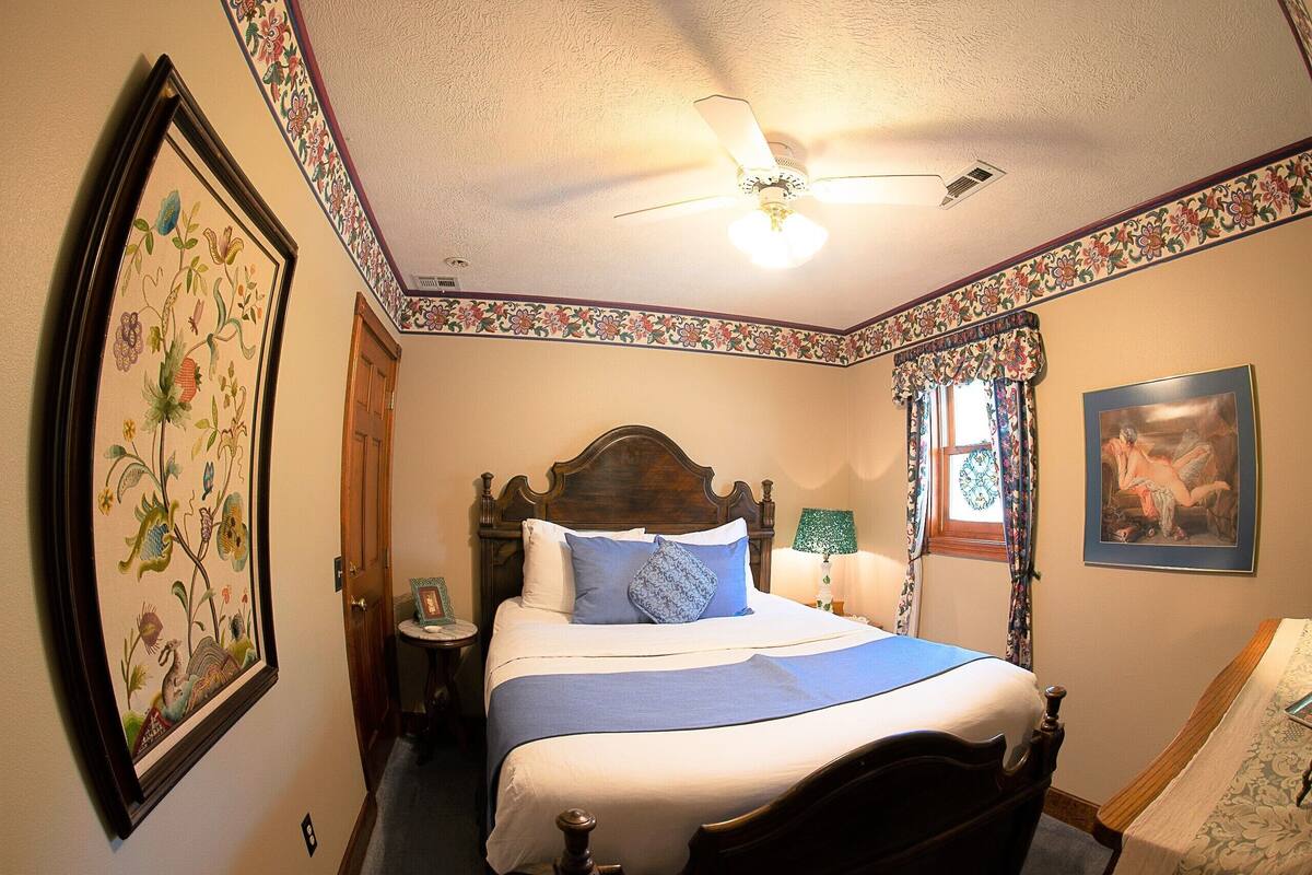 Un dormitorio con una cama de madera, un ventilador de techo, una obra de arte floral enmarco y una ventana con cortinas.