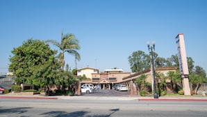 A hotel entrance with a parking lot, palm trees, and a clear blue sky. - Royala Inn (Montebello)