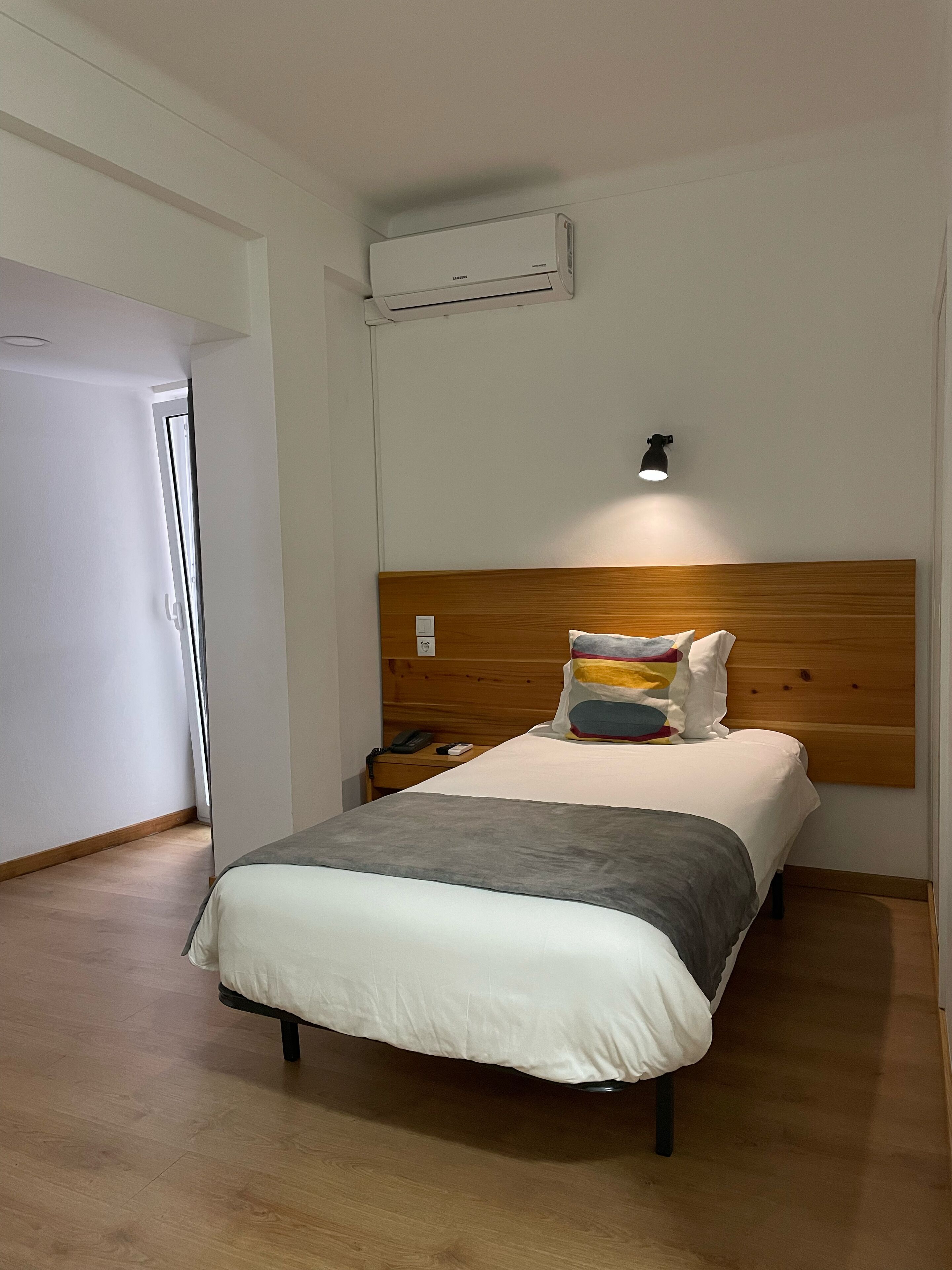 Una habitación de hotel con cabecera de madera, una cama con edredón gris y blanco, un teléfono en la mesita de noche y un equipo de aire acondicionado montado en la pared.