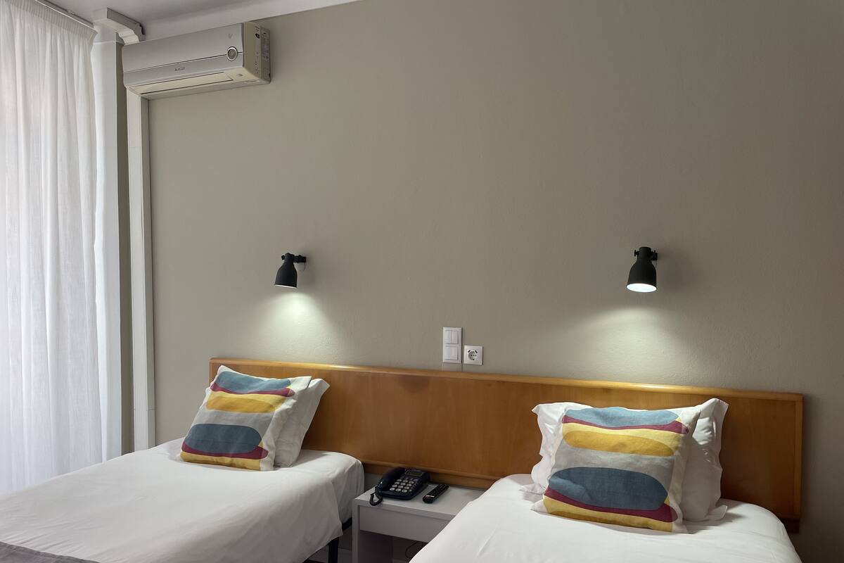 Ein Hotelzimmer mit zwei Betten, jeweils mit einem hölzernen Kopfteil und weißer Bettwäsche. Es gibt zwei Nachttische, einer mit Telefon und Fernbedienung, sowie zwei Wandlampen.
