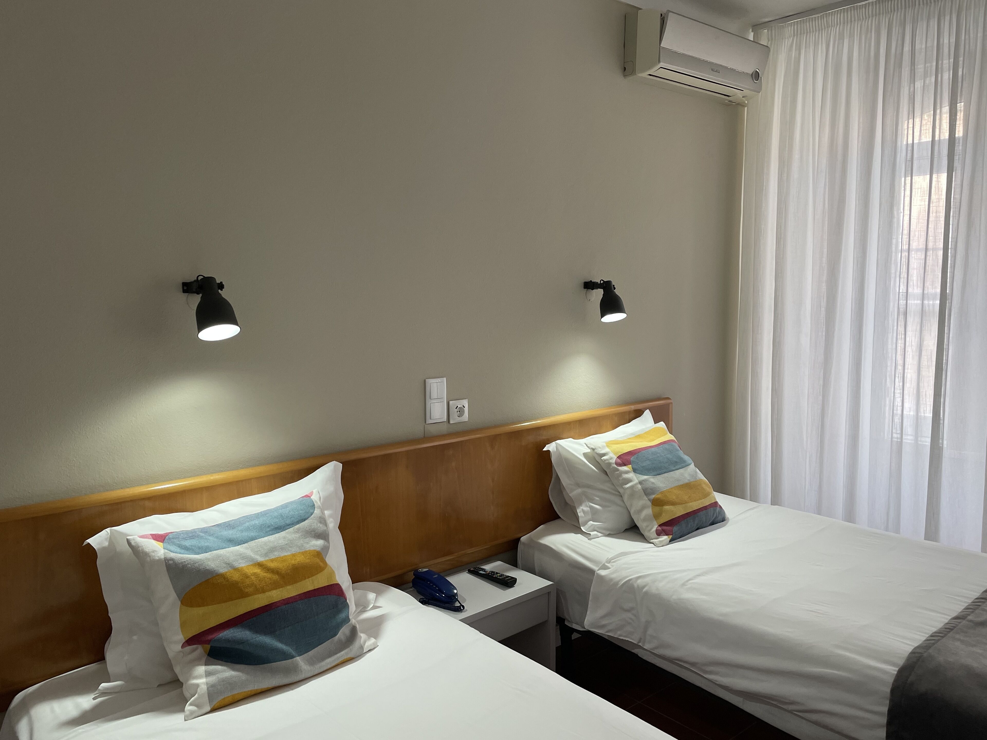 A hotel room with two beds, each with a striped pillow, a nightstand with a phone and a blue object, and two wall-mounted lights.
