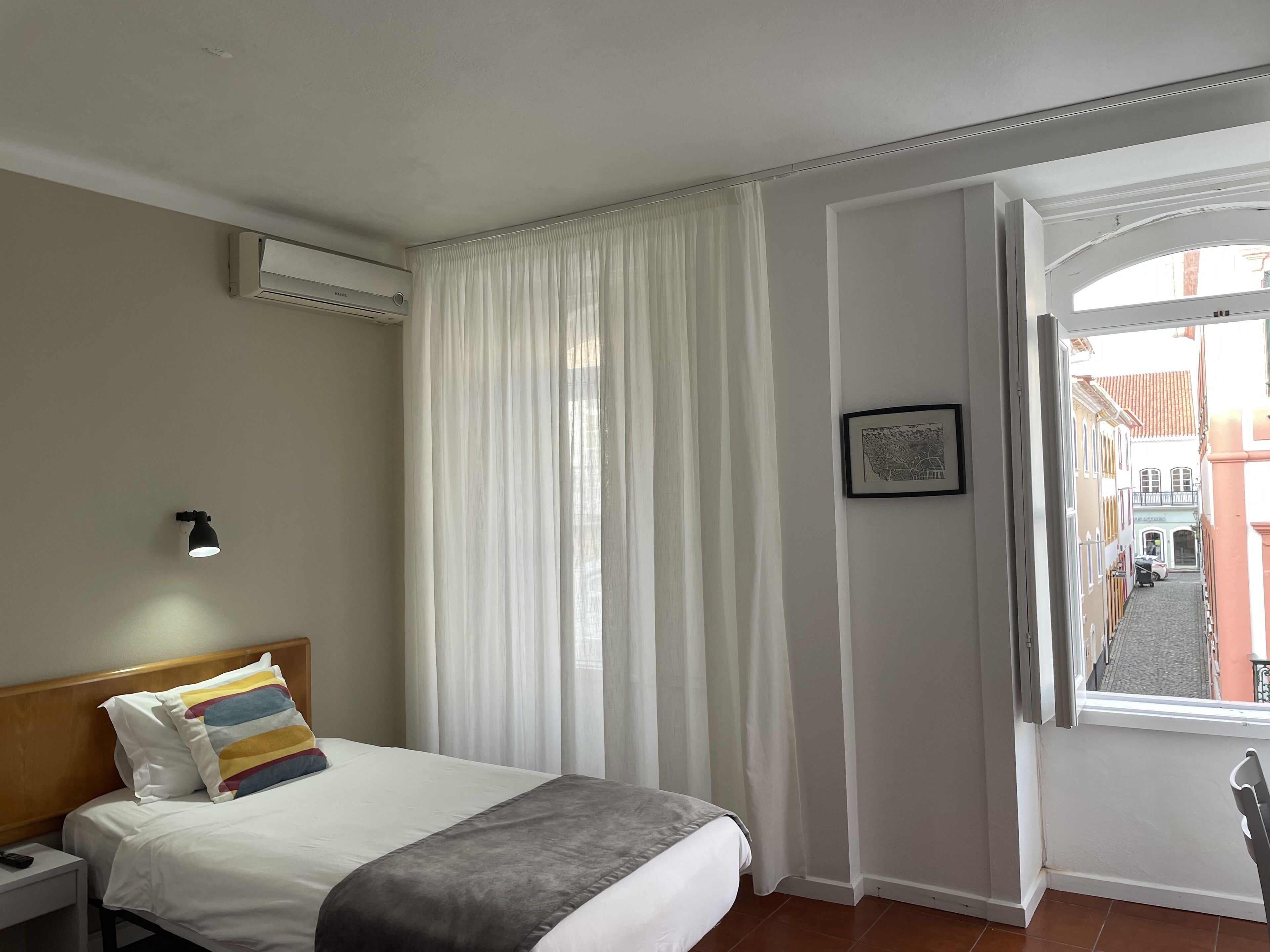 Una habitación de hotel con cama, mesita de noche, lámpara, ventana con cortinas y vista a la calle.