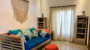 A cozy living room with a blue sofa, colorful cushions, a wooden side table, a red ottoman, a macrame wall hanging, a bookshelf, and a window with curtains. - Villa Legian (Legian)