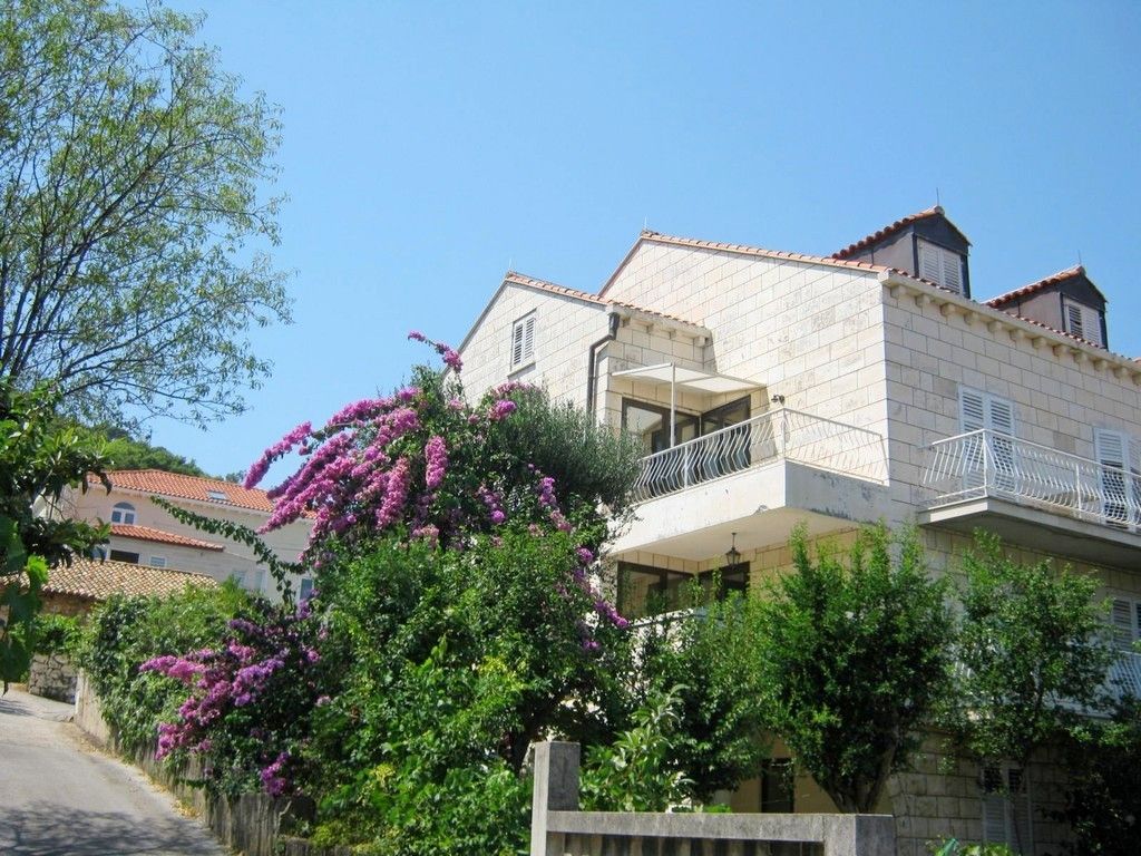 A two-story building with a balcony, surrounded by lush greenery and flowering plants.