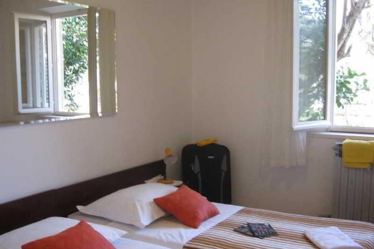 Una habitació d'hotel amb dos llits, una finestra que dona a arbres, un radiador i una maleta a terra.