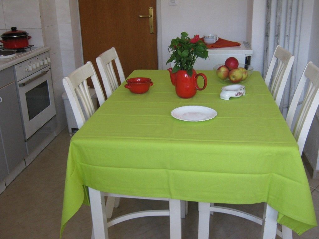 A dining table with a green tablecloth, white chairs, a red pitcher, a bowl of fruit, and a white plate.