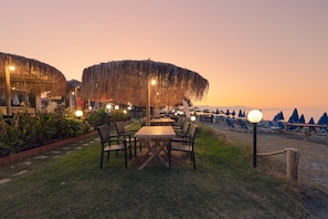An outdoor dining area with tables and chairs, surrounded by thatched umbrellas and potted plants. - Erato Beach Hotel (Platanias)