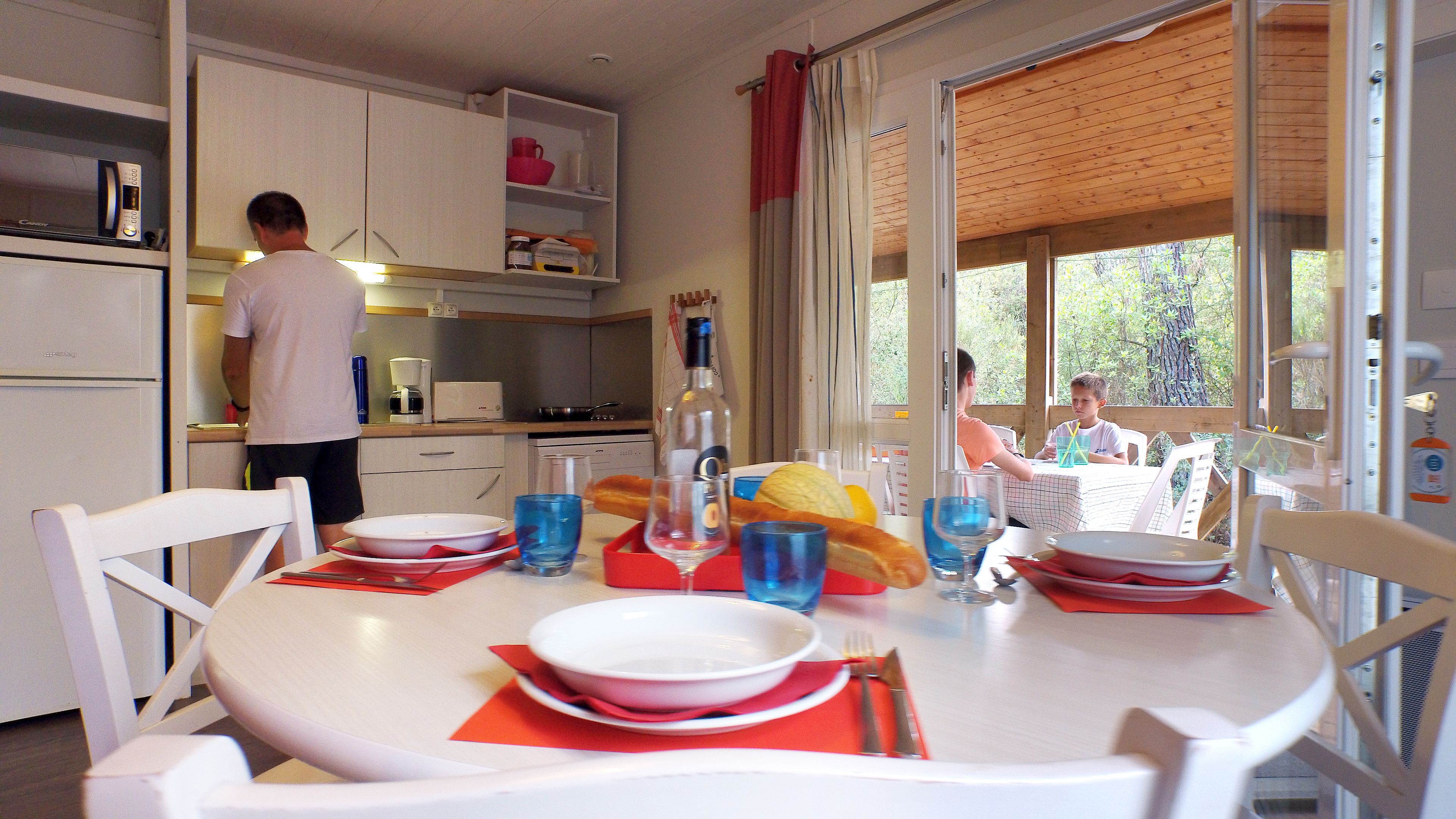 A dining area with a table set for two, a kitchen in the background, and a view of the outdoors.