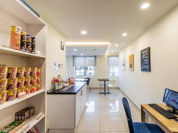 Una habitación de hotel con kitchenette, un comedor con mesa y sillas, un escritorio con teclado y monitor, y estantes llenos de aperitivos.