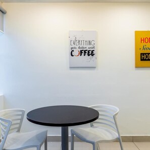 Ein Zimmer mit einem runden Tisch und vier Stühlen, eine weiße Wand mit zwei gerahmten Schildern, wobei eines 'EVERYTHING gets better with COFFEE' zeigt und das andere teilweise sichtbar ist.