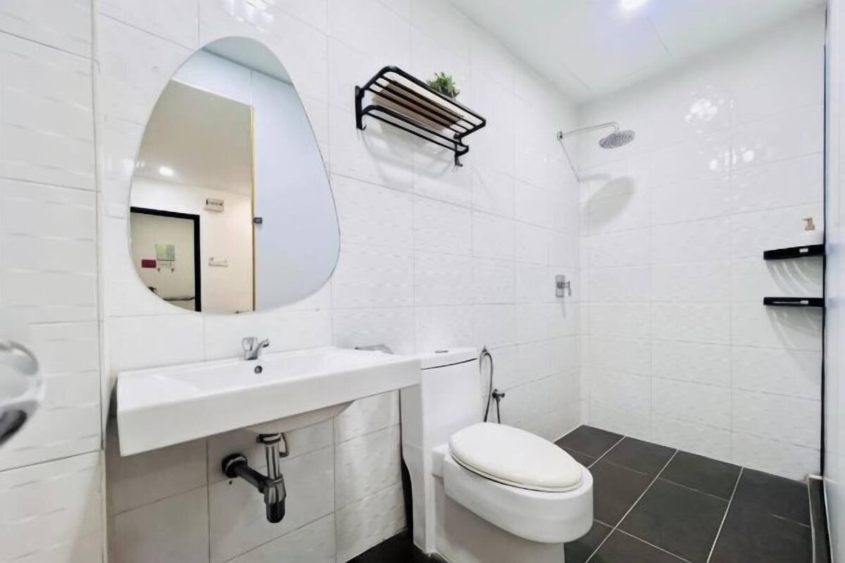 A bathroom with a white sink, toilet, and shower. There is a mirror above the sink and a shelf with a plant.