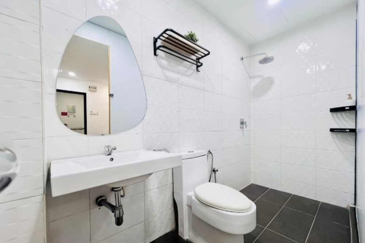 A bathroom with a white sink, toilet, and shower. There is a mirror above the sink and a shelf with a plant.