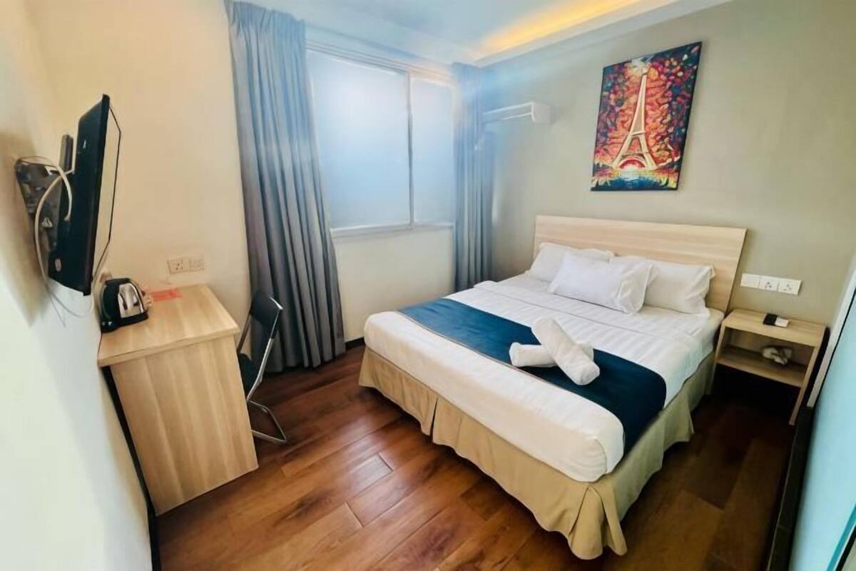 A hotel room with a bed, a desk, a chair, a TV, a window with curtains, and a painting of the Eiffel Tower.