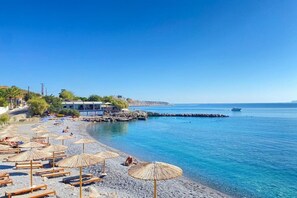 A beach with sun loungers and umbrellas, a building with a blue roof, and a clear blue sea. - Ariadne Beach (Hersonissos)