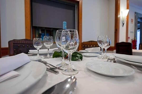 A dining table set with white plates, clear wine glasses, and silverware, with a television in the background. - Universal (Terras de Bouro)