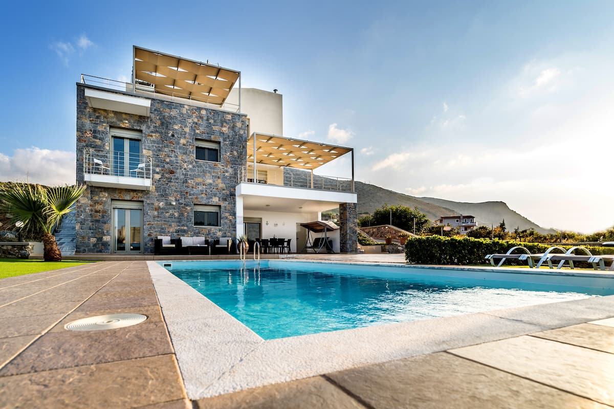 A modern two-story house with a swimming pool, stone facade, and a balcony with outdoor furniture.