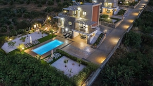 Una villa moderna de varios pisos con piscina, mobiliario exterior e iluminada entrada de vehículos.