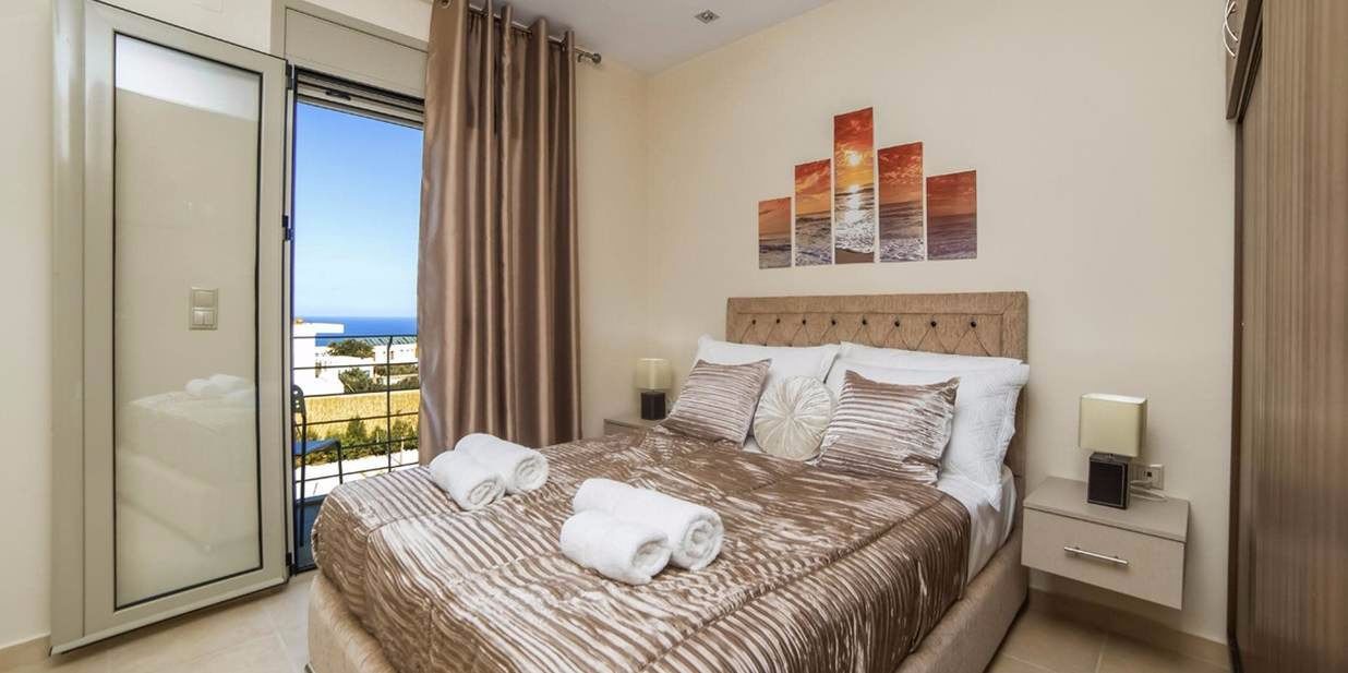A bedroom with a bed, a nightstand, a wall-mounted lamp, and a view of the sea through a sliding door.
