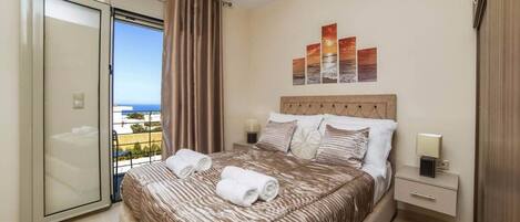 A bedroom with a bed, a nightstand, a wall-mounted lamp, and a view of the sea through a sliding door.