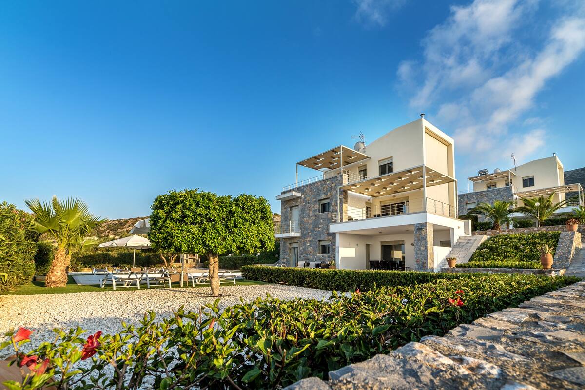 A modern two-story building with a stone facade, a balcony, and a well-maintained garden with a palm tree and shrubs.