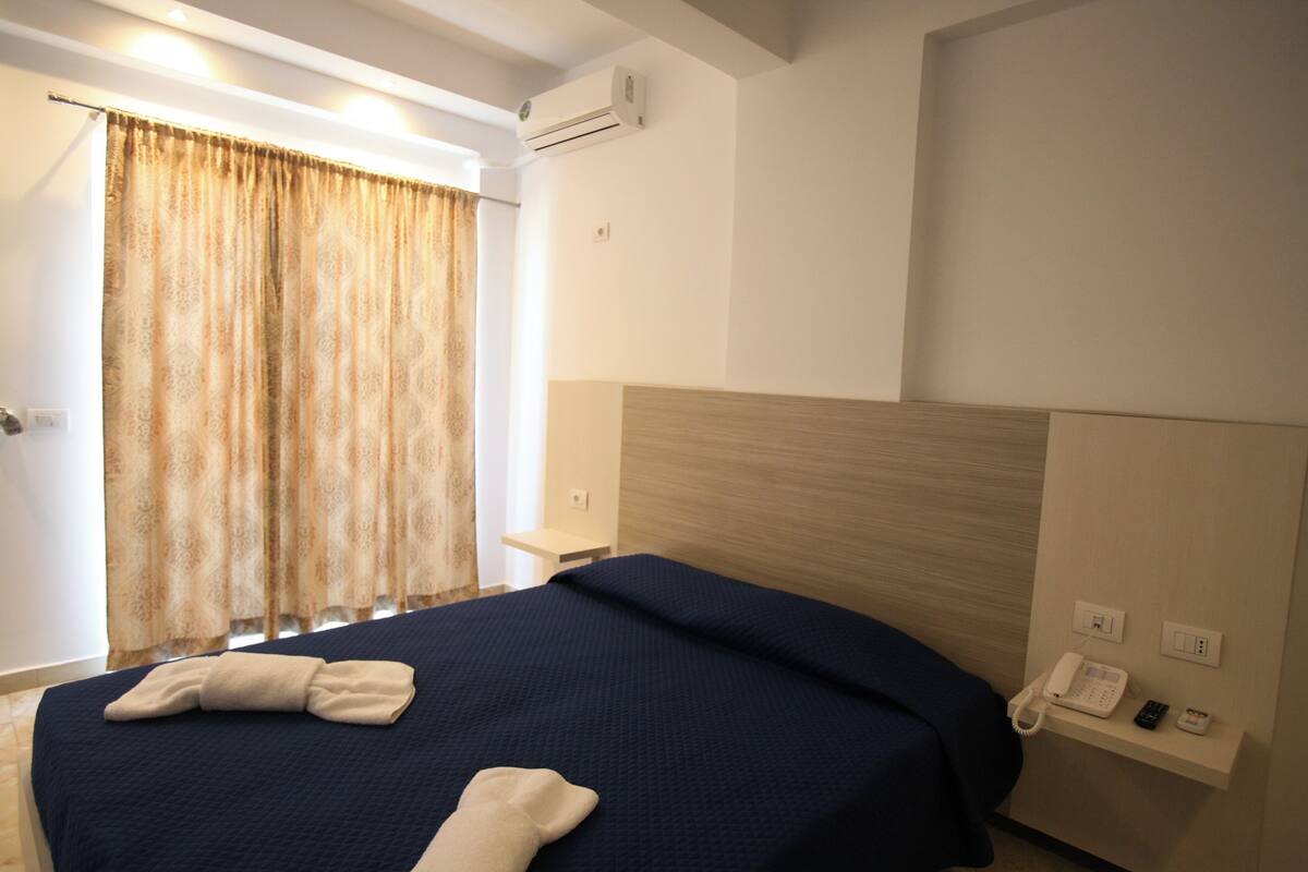 A hotel room with a large bed, a bedside table, an air conditioning unit, a telephone, and a curtain.