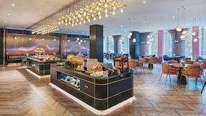 A modern hotel lobby with a central reception desk, seating areas, and a large window providing natural light.