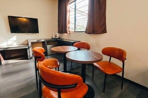 A room with a TV mounted on the wall, a seating area with round tables and orange chairs, and a window with curtains. - I Garden Hotel (Ipoh)