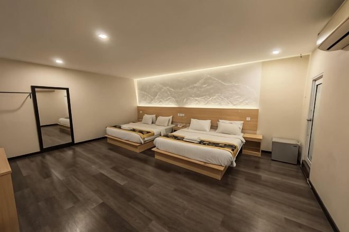 A hotel room with two beds, a wooden floor, a wall-mounted mirror, and a small bedside table.