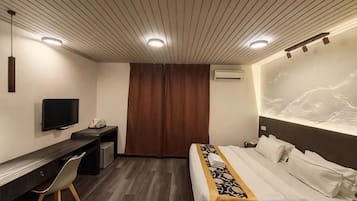 A hotel room with a bed, desk, chair, TV, and a wall-mounted air conditioner.