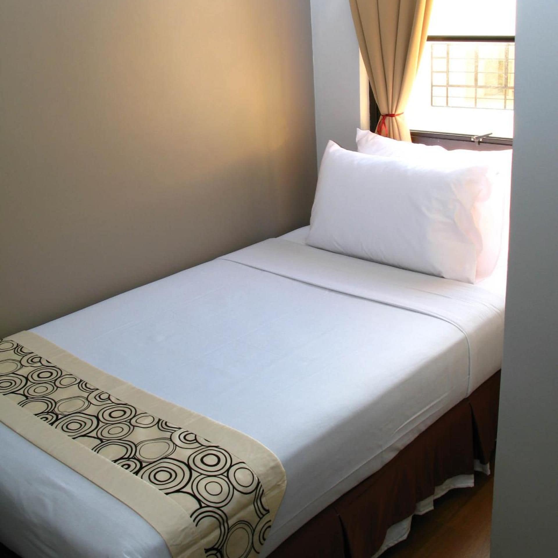 Ein Einzelbett mit einer gemusterten Tagesdecke, ein Fenster mit Vorhängen und eine schlichte Wand.