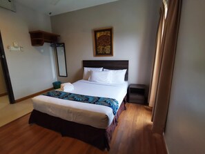 Una habitación de hotel con una cama grande, una mesita de noche, suelo de madera, un estante en la pared y un cuadro.