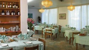 Un restaurant avec plusieurs tables, chacune recouverte d’une nappe vert clair. Des chaises en bois sont disposées autour des tables, et une étagère contenant des bouteilles de vin et des verres est visible au fond.